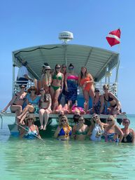 Group of women in colorful swimsuits posing on a covered party boat and in shallow turquoise water under a clear blue sky with a red diver-down flag.