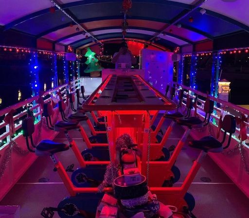 Nighttime holiday-lit party pedal boat interior with rows of bike-style seats and pedals around a central bar, colorful string lights and an inflatable Christmas tree reflecting on nearby water.
