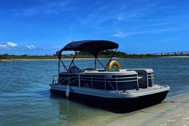 Pontoon boat beached on a sandy shore in a calm coastal inlet, bright blue sky overhead, golden inflatable tube onboard, with marshland and a low bridge in the distance.