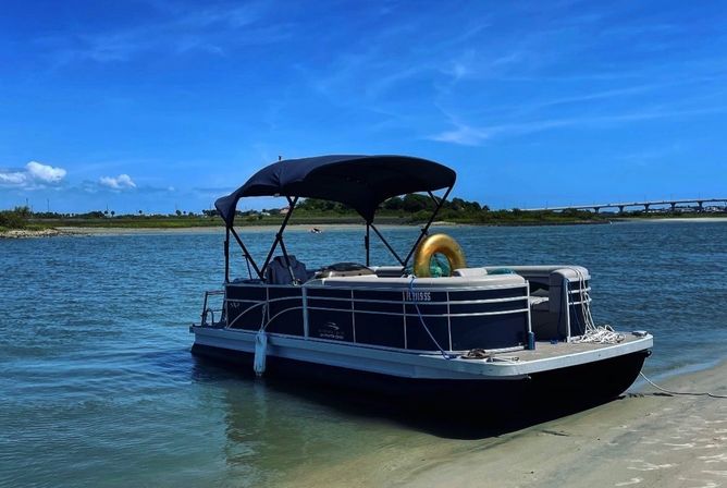 Pontoon boat beached on a sandy shore in a calm coastal inlet, bright blue sky overhead, golden inflatable tube onboard, with marshland and a low bridge in the distance.