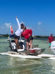 Two women striking a joyful pose on a small pontoon-style boat over clear shallow turquoise water at a sunny sandbar; one in a white long-sleeve and cap raises an arm, the other in a red top and sunglasses smiles, with a man wading nearby and coastal homes on the distant shoreline.