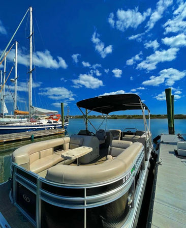 Cream-colored pontoon boat moored at a sunny marina dock beside sailboats, calm reflective water and a vivid blue sky dotted with fluffy white clouds, perfect waterfront scene.