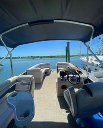 Sunny pontoon boat interior with helm, cushioned lounge seating and cup holders under a bimini, looking out at a calm marina with dock pilings and sailboats.