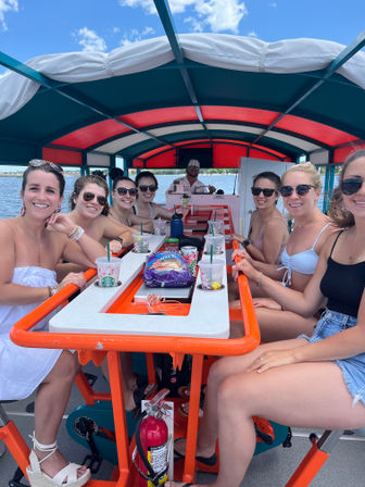 Smiling group of friends on a covered orange-and-teal pedal pub boat on a sunny lake, seated around a central drink-and-snack table with blue sky and shoreline visible.