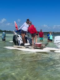 Two women smiling and posing on a small motorized pontoon at a shallow sandbar in clear turquoise water under a bright blue sky, with other people wading and taking photos in the background