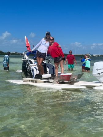 Two women smiling and posing on a small motorized pontoon at a shallow sandbar in clear turquoise water under a bright blue sky, with other people wading and taking photos in the background