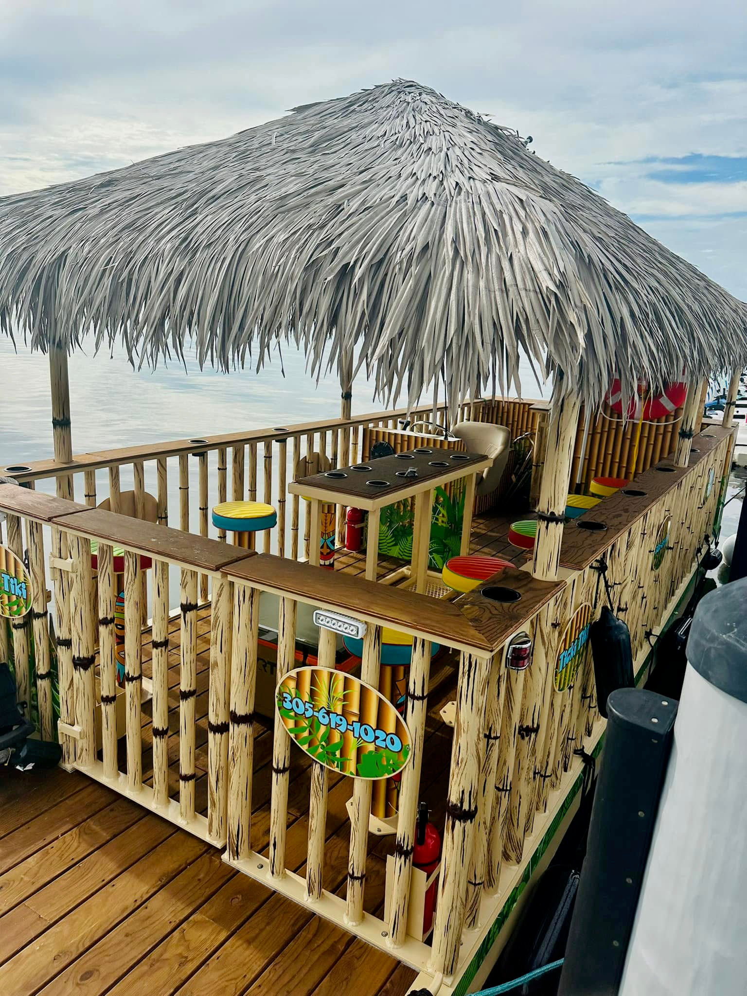 Floating tiki bar with palm-thatched roof, bamboo railings and colorful round stools docked at a calm marina waterfront