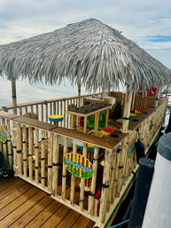 Floating tiki bar with palm-thatched roof, bamboo railings and colorful round stools docked at a calm marina waterfront
