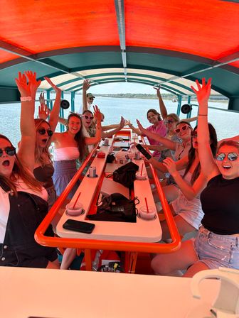 Group of friends cheering with hands raised on a covered party boat over calm coastal waters, bright red canopy and central picnic table with drinks — sunny summer outing.