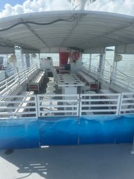 Shaded pontoon-style boat deck with long bench seating, stools and a central table with cup/rod holders, life ring on the stern, blue wave panels and calm coastal waters under a partly cloudy sky.