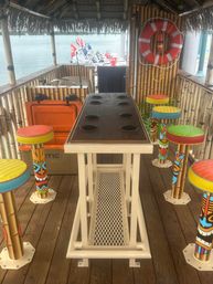 Thatched-roof tiki hut on a wooden dock overlooking calm water, featuring a long cup-holder table, colorful painted tiki bar stools, bamboo walls, a life preserver, and boats moored in the background.