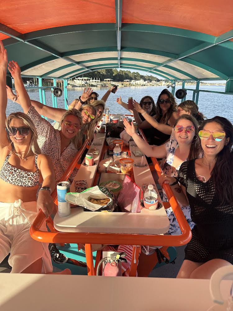 Group of friends on a covered party boat at a sunny lake — women in colorful sunglasses smiling and waving around a central table with snacks, drinks and a bright canopy with shoreline docks in the background