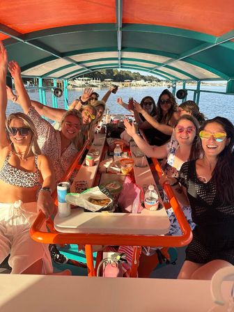 Group of friends on a covered party boat at a sunny lake — women in colorful sunglasses smiling and waving around a central table with snacks, drinks and a bright canopy with shoreline docks in the background