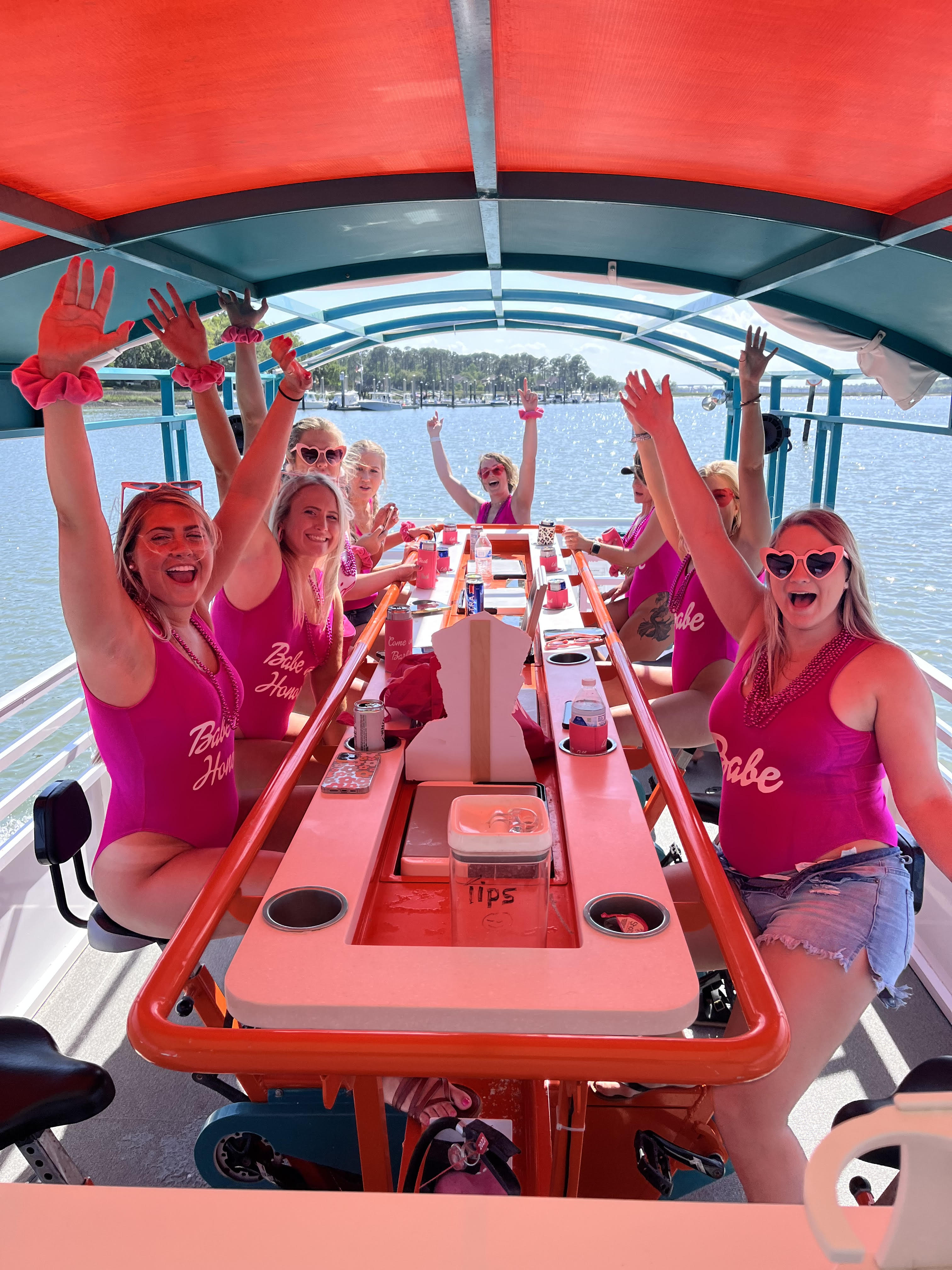 Group of women in matching pink swimsuits and heart sunglasses cheering on a colorful pedal party boat at a sunny marina — bachelorette-style waterfront celebration