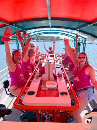 Group of women in matching pink swimsuits and heart sunglasses cheering on a colorful pedal party boat at a sunny marina — bachelorette-style waterfront celebration