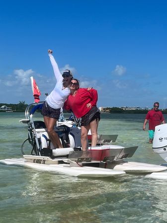 Two people posing and smiling on a twin-hull pontoon boat anchored in shallow clear turquoise coastal water by a sandbar, bright blue sky, red flag on the stern and shoreline buildings in the distance.