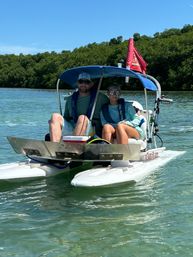 Two people in sunglasses and life jackets seated under a blue canopy on a compact pontoon-style boat cruising clear green water past a mangrove-lined coastal shoreline on a sunny day.