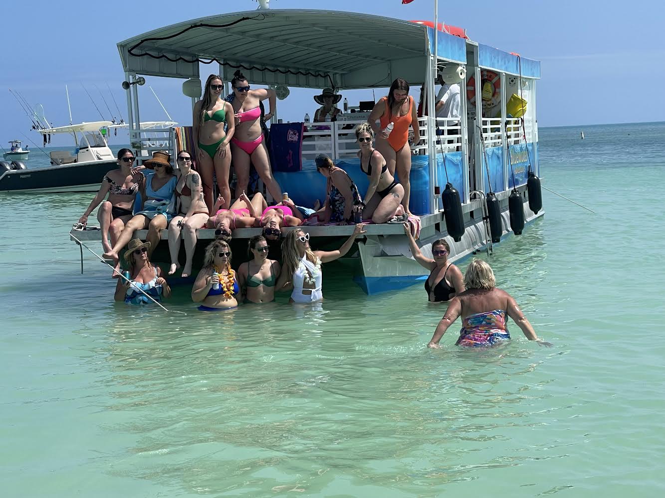 Group of women enjoying a sunny boat party on a colorful pontoon, lounging on the deck and wading in shallow turquoise ocean near a tropical beach.