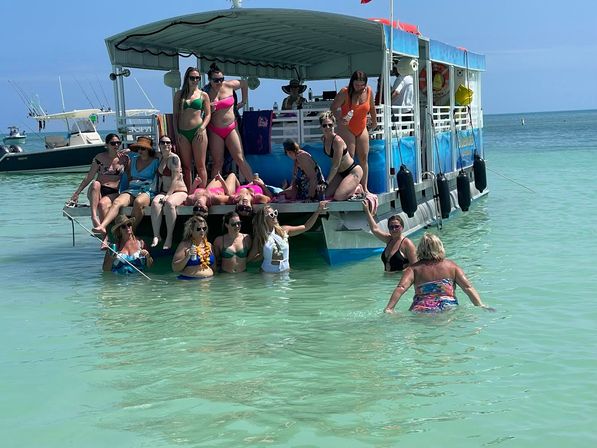 Group of women enjoying a sunny boat party on a colorful pontoon, lounging on the deck and wading in shallow turquoise ocean near a tropical beach.