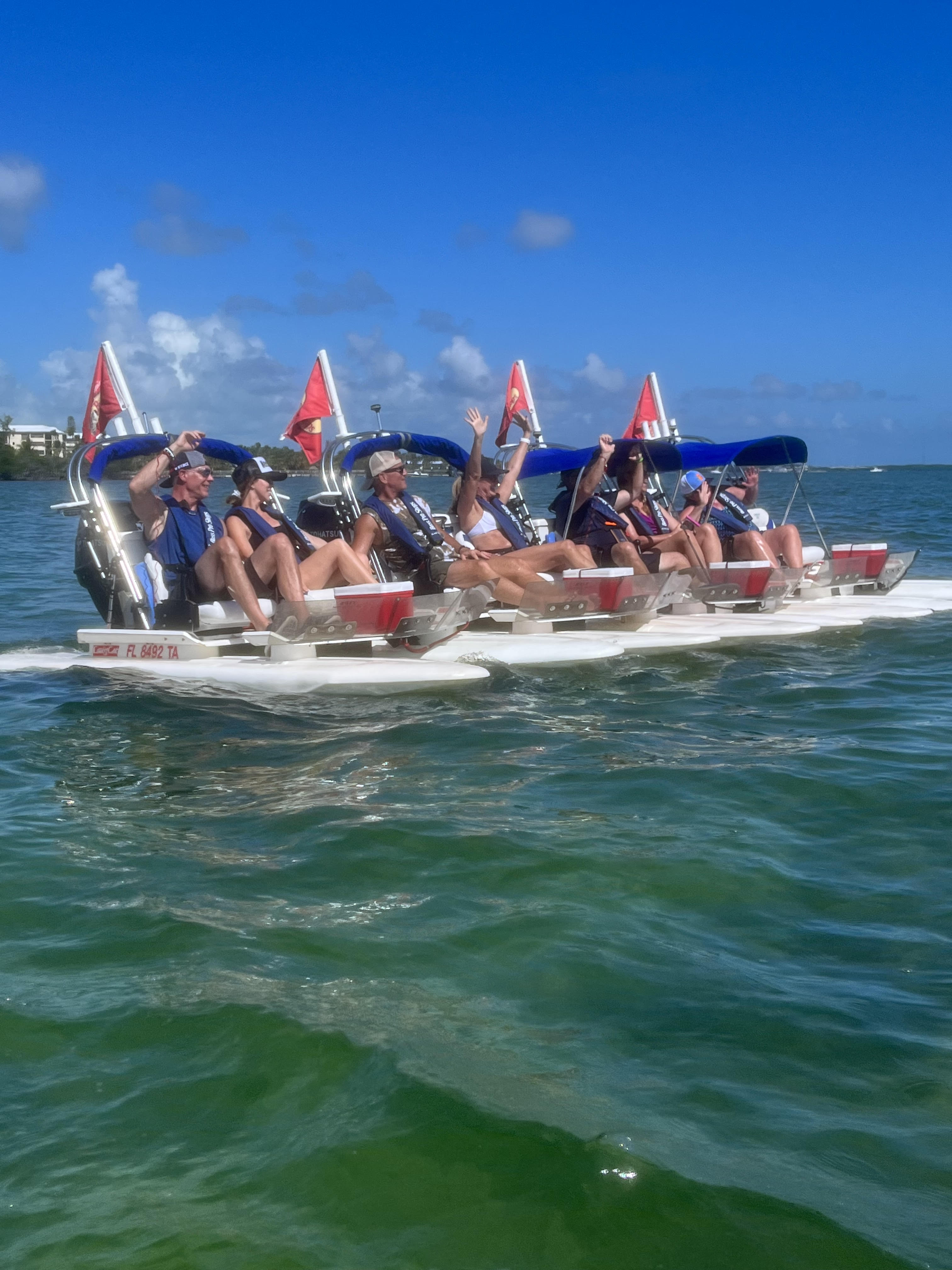 Group of adults in life jackets seated on connected white pontoon pedal boats with blue canopies and red dive flags, waving while cruising turquoise coastal water under a bright blue sky.