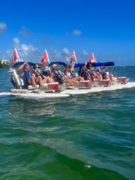Group of adults in life jackets seated on connected white pontoon pedal boats with blue canopies and red dive flags, waving while cruising turquoise coastal water under a bright blue sky.
