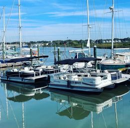 Sunny coastal marina with pontoon boats and sailboats docked, black canvas canopies and tall masts reflected in calm blue-green harbor water.