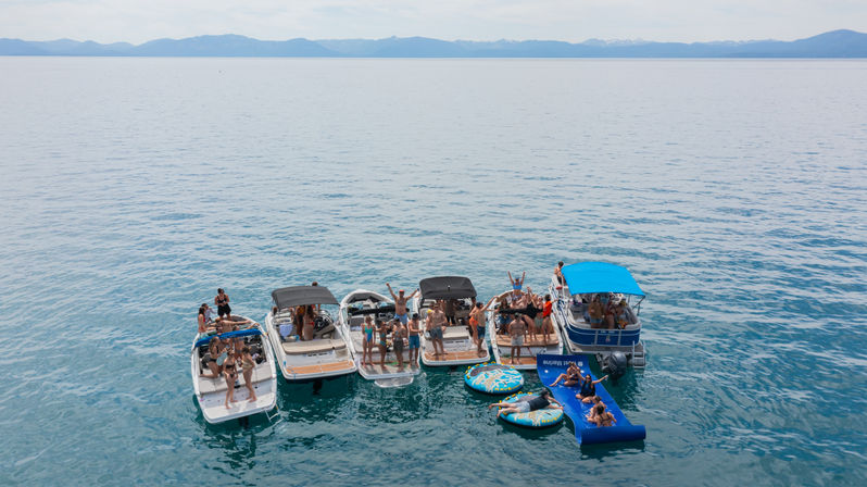 Aerial view of a summer boat party: five motorboats rafted together on a clear mountain lake, people socializing on decks and colorful inflatables and a floating mat in the water with a distant mountain range on the horizon.