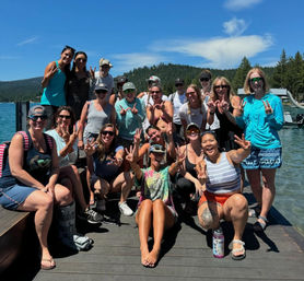 Cheerful group posing on a wooden dock at a sunny mountain lake with pine-covered hills, smiling and flashing peace signs — summer lakeside and boating recreation.