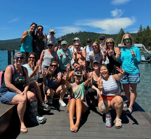Cheerful group posing on a wooden dock at a sunny mountain lake with pine-covered hills, smiling and flashing peace signs — summer lakeside and boating recreation.