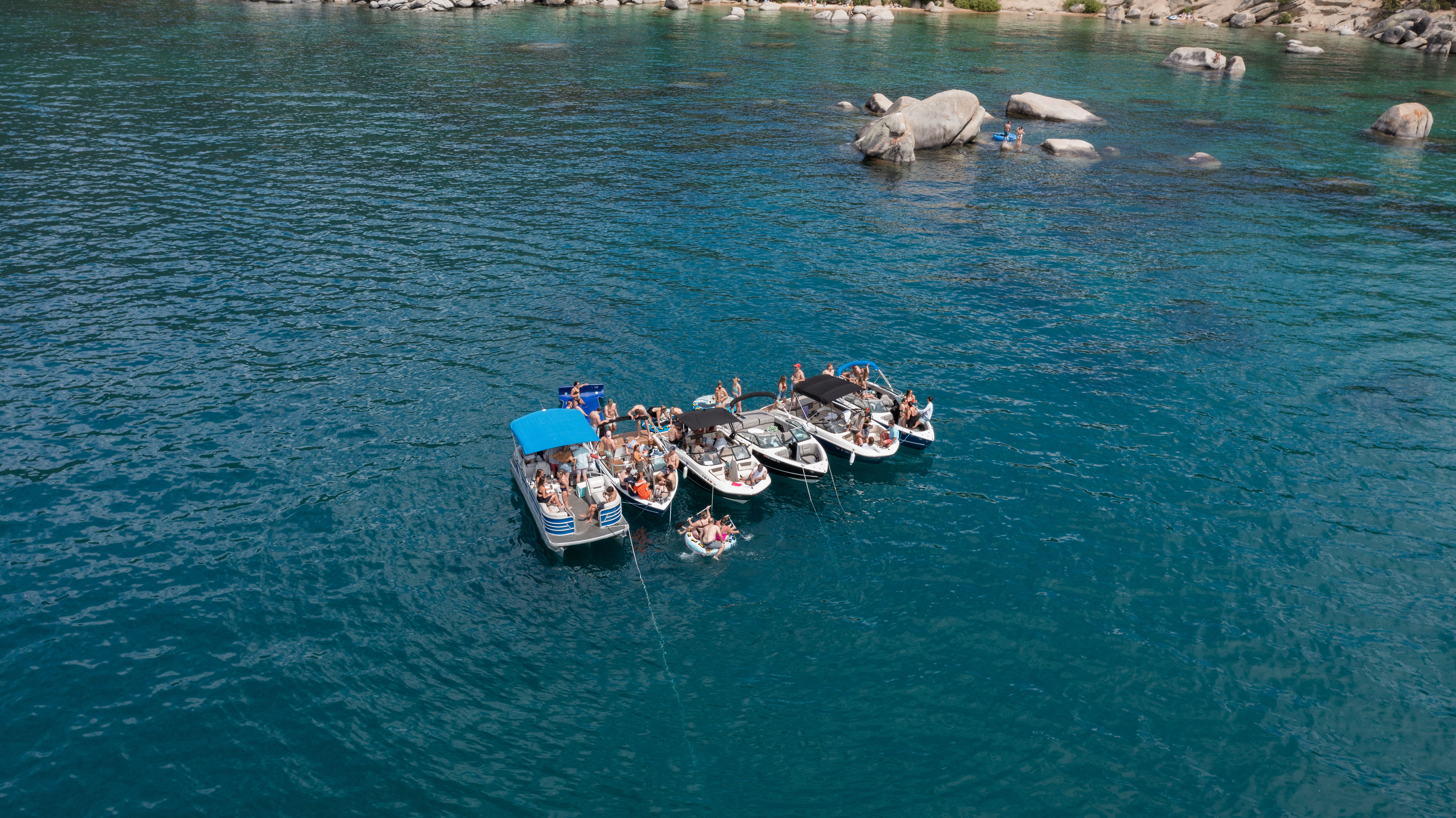 Aerial shot of five motorboats rafted together with people sunbathing and floating on a clear turquoise lake near a rocky shoreline and boulders on a sunny summer day.