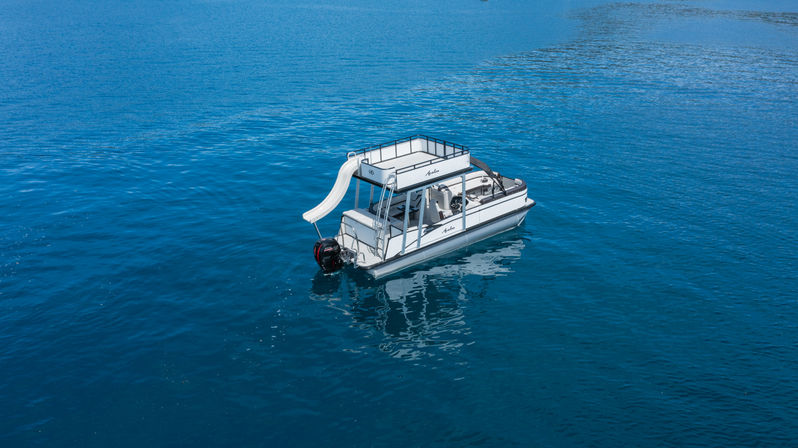 Aerial view of a white pontoon boat with rooftop deck and water slide drifting on calm deep-blue sea