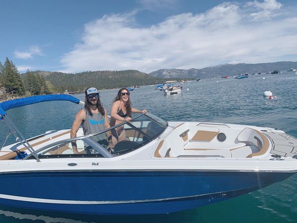 Two people laughing on a blue-and-white motorboat on a sunny mountain lake, with anchored boats and a forested shoreline in the background.