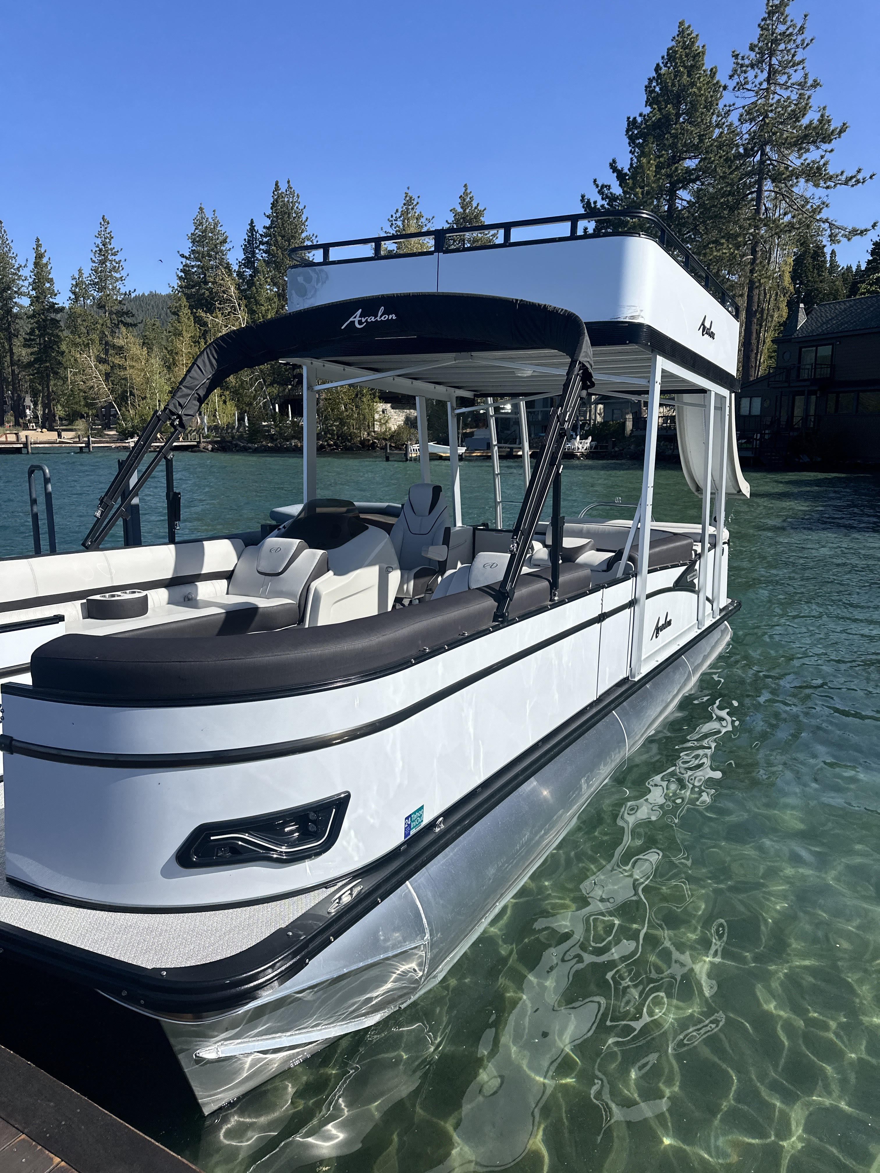Sleek white double-deck pontoon boat docked on a crystal-clear mountain lake with pine trees and lakeside cabins under a bright blue sky