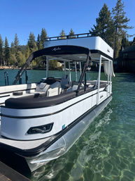 Sleek white double-deck pontoon boat docked on a crystal-clear mountain lake with pine trees and lakeside cabins under a bright blue sky