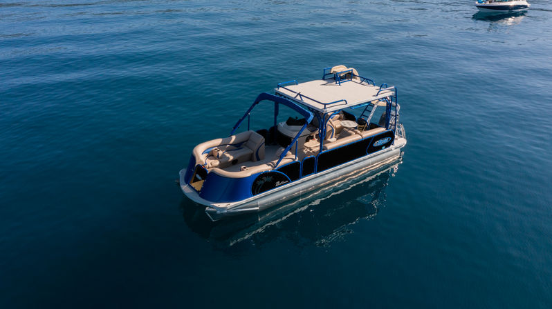 Aerial view of a blue-and-tan pontoon boat with shaded canopy and cushioned seating floating on calm deep-blue water on a sunny day