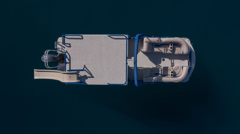 Aerial top-down view of a pontoon boat with slide, cushioned lounge seating and outboard motor floating on calm deep-blue lake water.