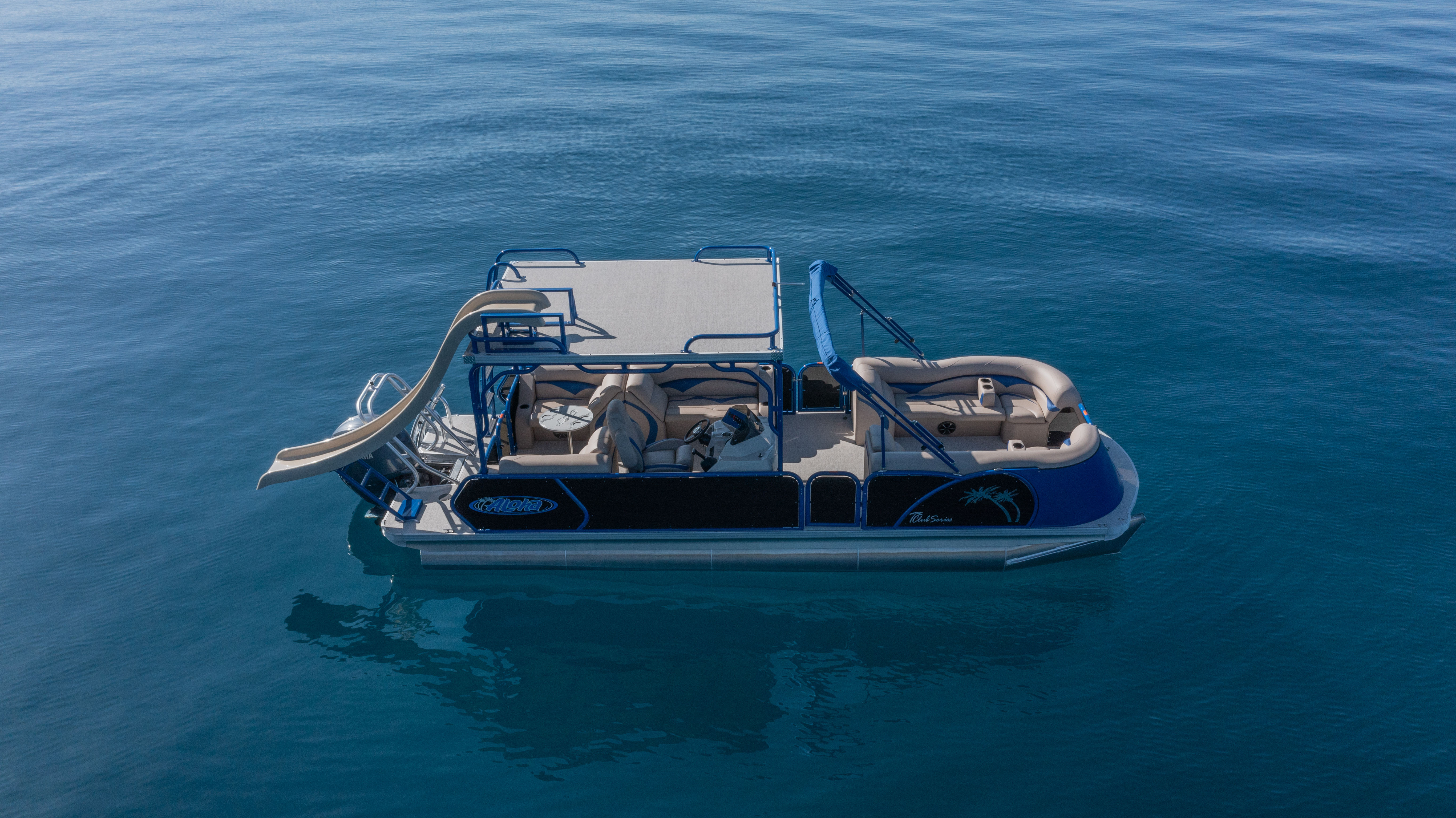Aerial view of a blue-and-beige pontoon boat with a deck and waterslide floating on calm deep-blue water.