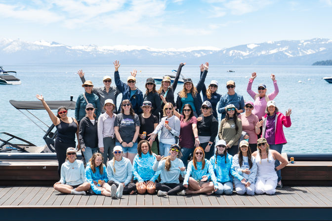 Cheerful large group of people in casual outdoor gear smiling and waving on a wooden lakeside dock, with boats on the water and snow-capped mountains under a bright blue sky.