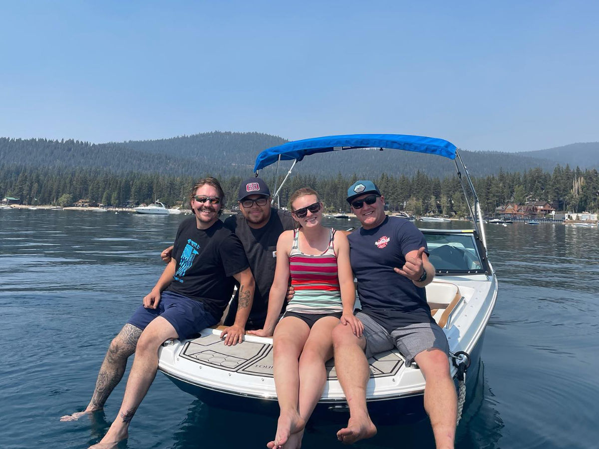 Four friends sitting on the bow of a recreational boat on a calm mountain lake with pine-forested shoreline and blue canopy, smiling on a sunny day