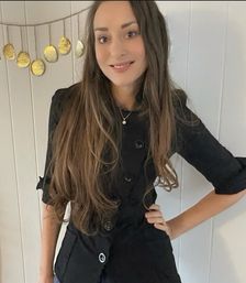 Smiling woman in a black buttoned jacket with long brown hair posed indoors against white paneled wall and a gold disc garland — casual indoor portrait.