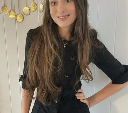 Smiling woman with long brown hair wearing a black buttoned jacket, posing indoors against a white paneled wall with hanging gold disc decorations.