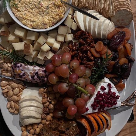 Overhead shot of a gourmet charcuterie board with cubed and sliced cheeses, brie topped with pomegranate seeds, a grape cluster, candied nuts, dried apricots and dates, citrus slices, crackers and a creamy dip — a festive party appetizer platter.