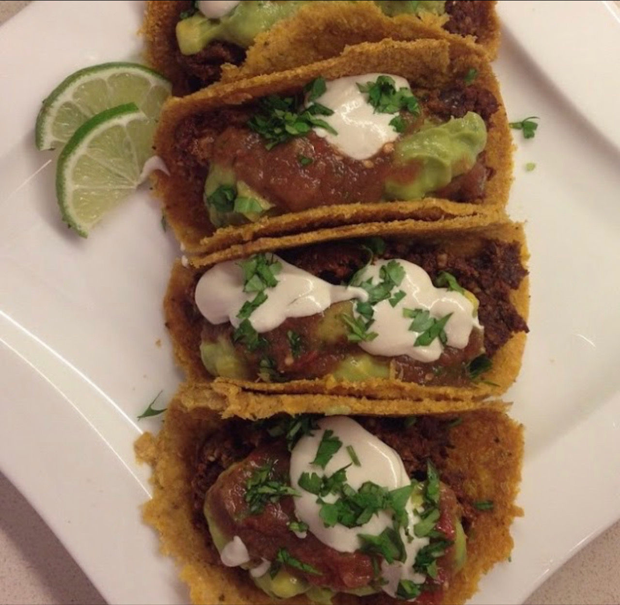 Four crispy golden corn-shell tacos filled with seasoned beef, guacamole, red salsa and sour cream, garnished with chopped cilantro and lime wedges on a white plate