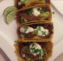 Four crispy golden corn-shell tacos filled with seasoned beef, guacamole, red salsa and sour cream, garnished with chopped cilantro and lime wedges on a white plate
