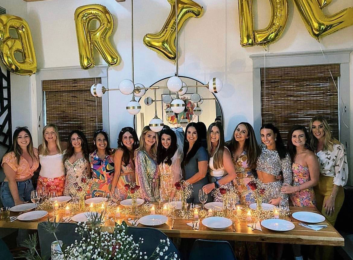 Bachelorette party dinner with a smiling group of women behind a candlelit wooden table, gold 'BRIDE' balloons, round mirror and modern pendant light.