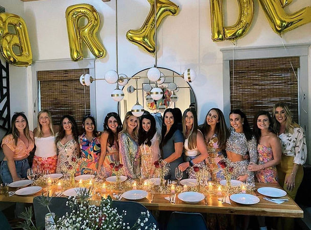 Bachelorette party dinner with a smiling group of women behind a candlelit wooden table, gold 'BRIDE' balloons, round mirror and modern pendant light.