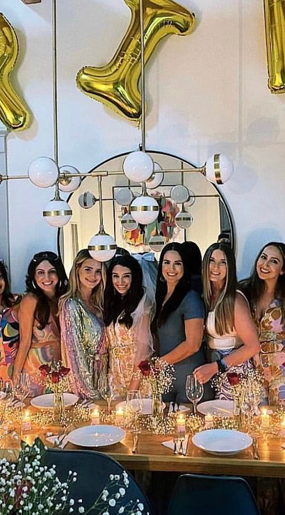 Bachelorette party dinner with a smiling group of women behind a candlelit wooden table, gold 'BRIDE' balloons, round mirror and modern pendant light.