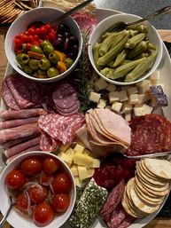 Colorful charcuterie grazing board with assorted cured meats (salami, prosciutto, turkey), cubed cheeses, herb-coated goat cheese log, bowls of olives, pickled okra, marinated cherry tomatoes, crackers and cranberry chutney — perfect party appetizer platter.