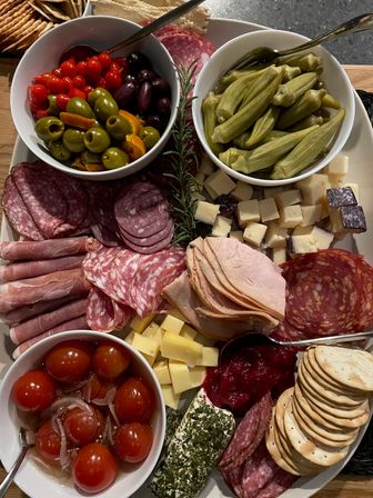 Colorful charcuterie board with marinated olives, pickled okra, cherry tomatoes, assorted salami and ham, cubed cheeses, herb-crusted log, crackers and rosemary garnish.