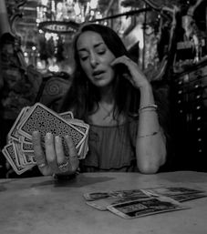 Black-and-white photo of a tarot reader shuffling a deck over a table with a spread of tarot cards, ornate chandelier and vintage parlor decor in the background.